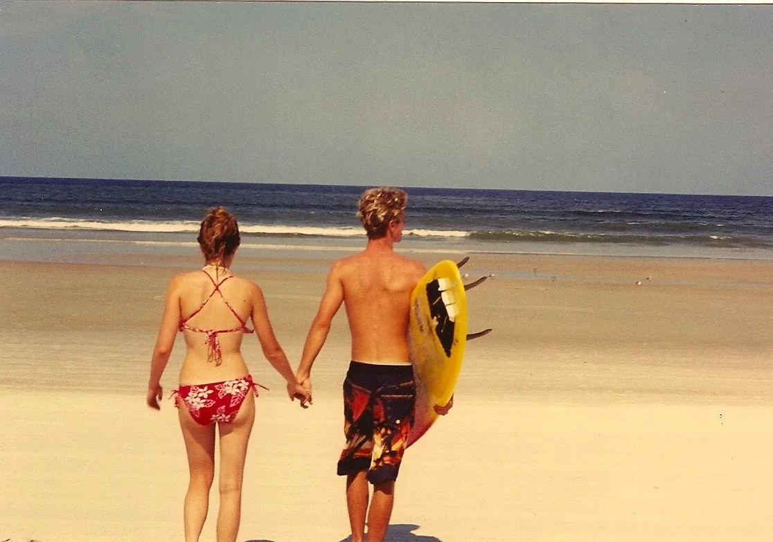 Young Daniel and Steph walking hand in hand toward the ocean, Daniel carrying a yellow surfboard